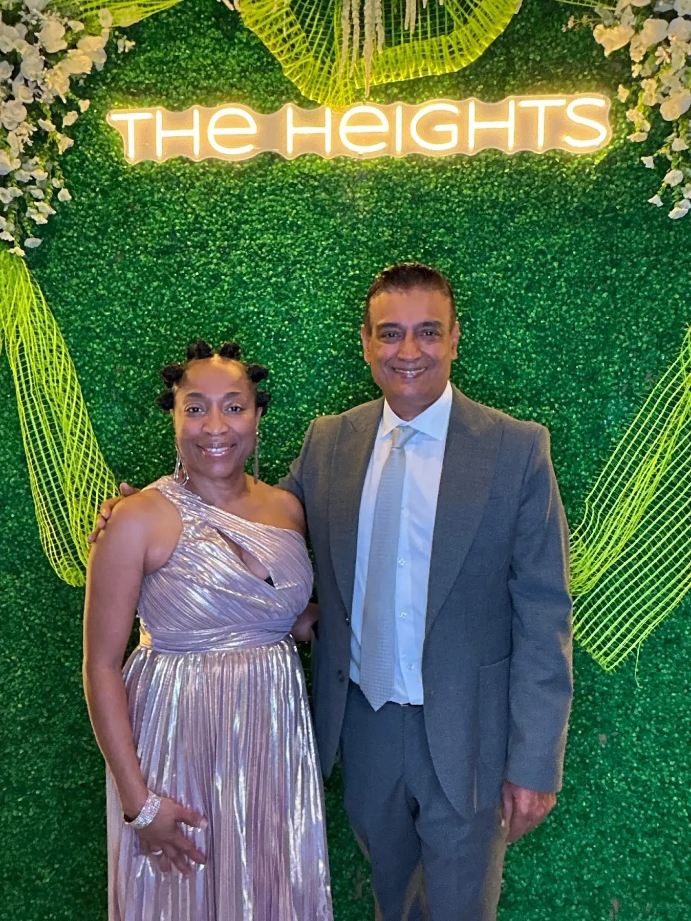 A couple in front of the neon The Heights sign on the greenery wall