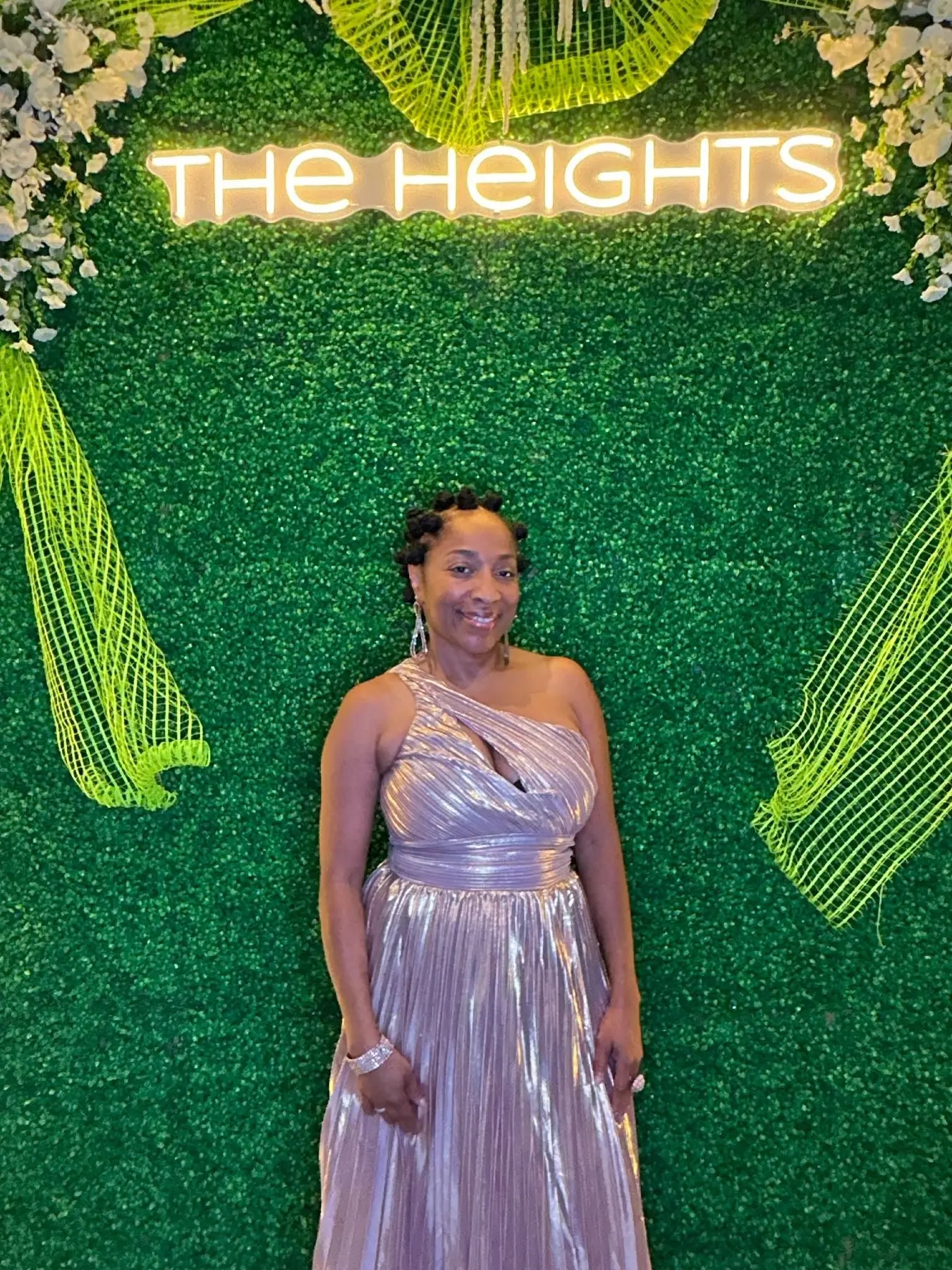 Guest in a champagne gown in front of the neon The Heights sign on the greenery wall